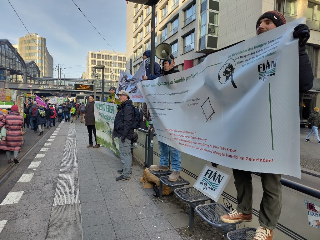 Auf dem Bild ist eine Straße mit Stadtbahnhaltestelle zu sehen. Am linken Bildrand schlängelt sich ein Demonstrationszug entland und rechts stehen Aktivist*innen von FIAN und anderen Organisationen mit Bannern und einem Megafon.